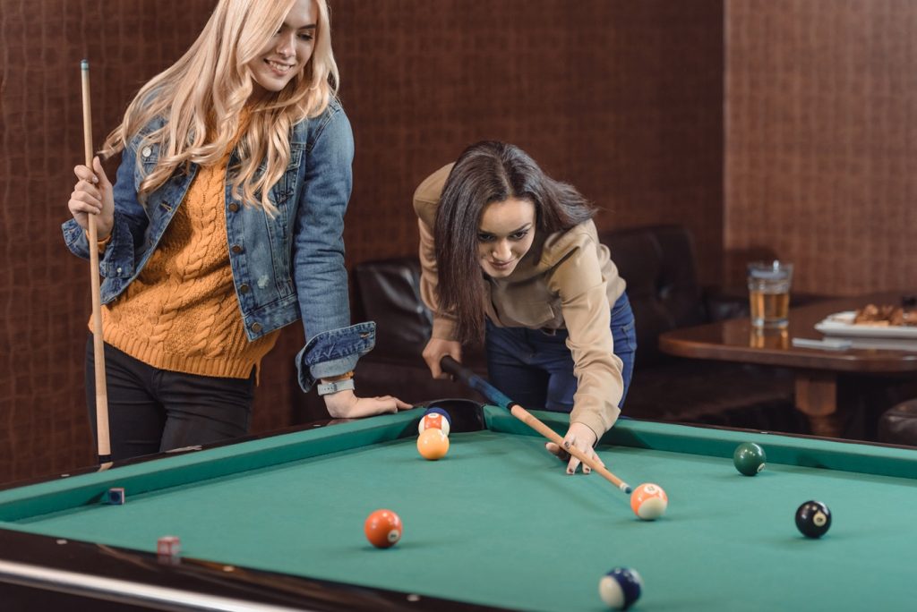 two women friends playing pool at a bar waiting for a man to approach them and talk to them