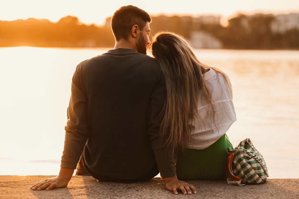 A couple sitting and enjoying sunset near a city lake, being physically intimate and comfortable with each other