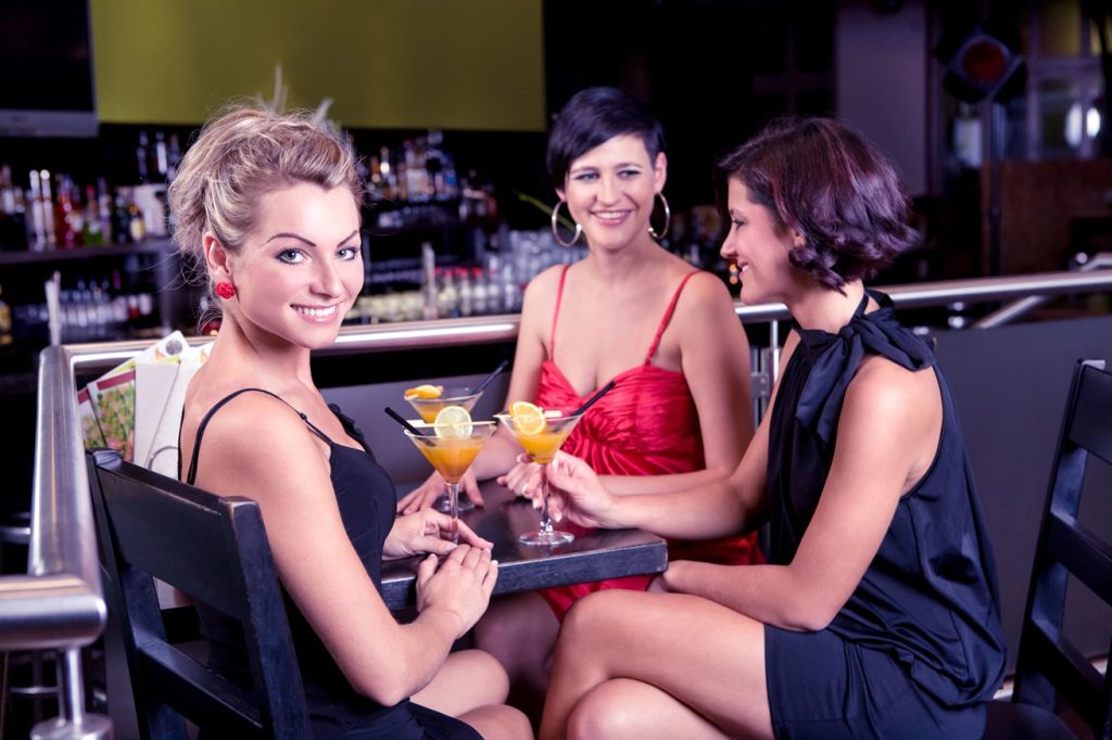 Three women sitting in a bad, having drinks, talking to each other, one of them looking at you
