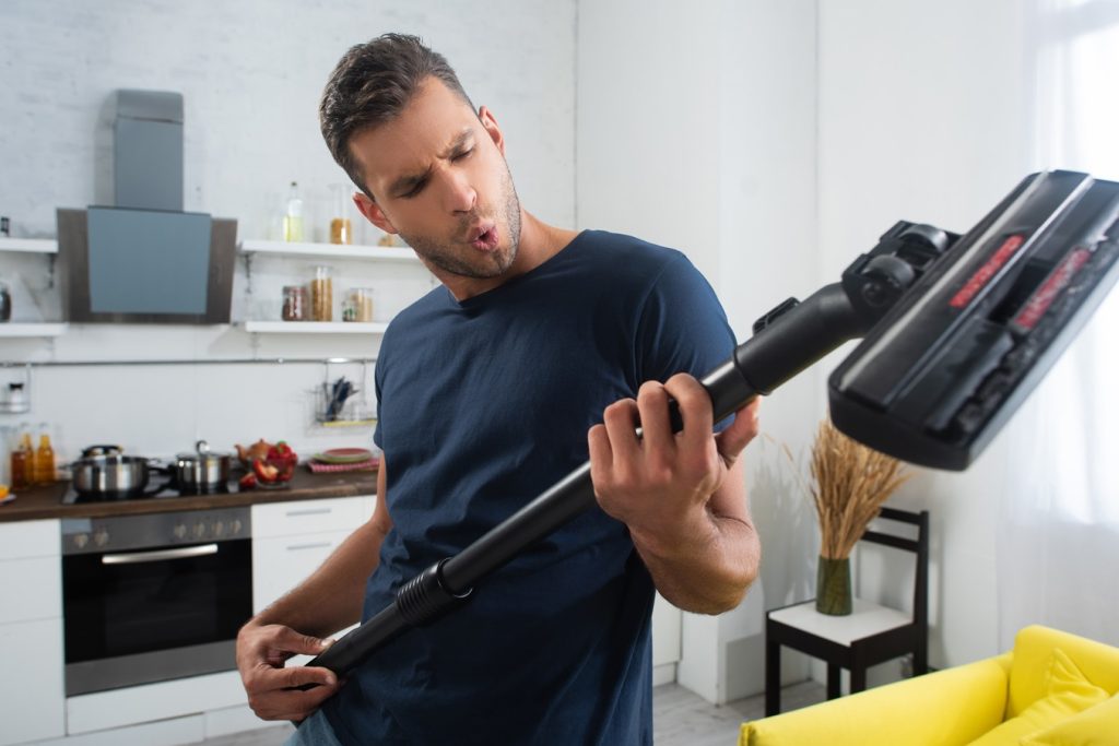 Excited man full of self-amusement having fun with brush of vacuum cleaner at home