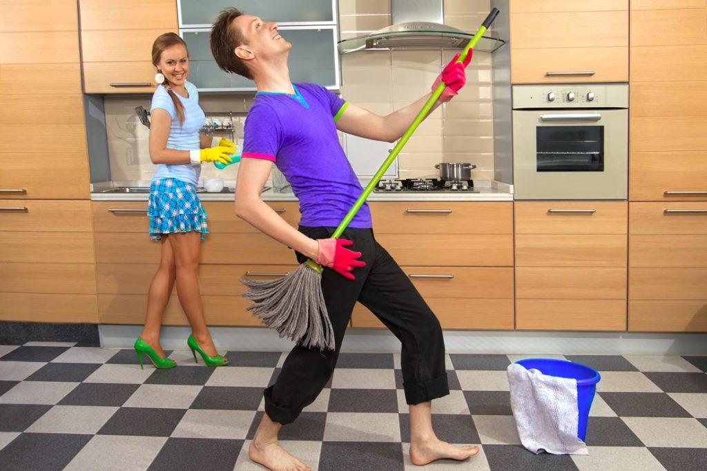 woman having fun with man who is amusing himself with a broom, pretending it's a guitar, while doing chores, showing that it's easy to seduce women with your personality
