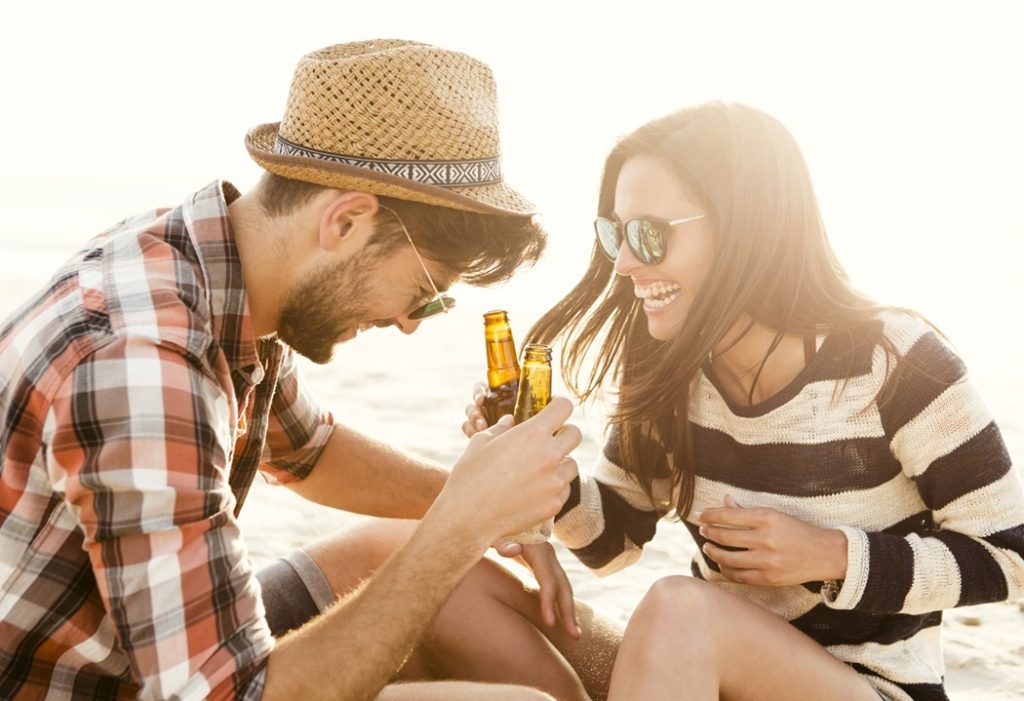 Young couple at the beach having fun, laughing and drinking beer, discussing how to seduce women with your personality